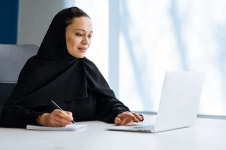 Beautiful woman with abaya dress working on her computer. Middle aged female employee at work in a business office in Dubai. Concept about middle eastern cultures and lifestyleの写真素材