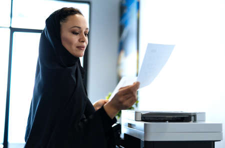 beautiful woman with abaya dress working and printing documents. Middle aged female employee at work in a business office in Dubai. Concept about middle eastern cultures and lifestyleの写真素材
