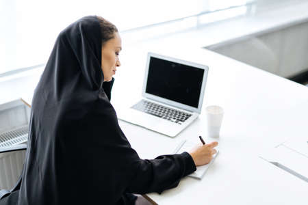 Beautiful woman with abaya dress working on her computer. Middle aged female employee at work in a business office in Dubai. Concept about middle eastern cultures and lifestyleの写真素材
