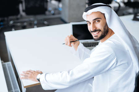 Handsome man with dish dasha working in his business office of Dubai. Portraits of a successful businessman in traditional emirates white dress. Concept about middle eastern cultures, lifestyle and professional occupationsの写真素材