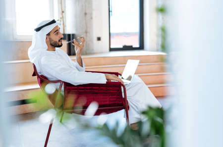 handsome man with dish dasha working in his business office of Dubai. Portraits of a successful businessman in traditional emirates white dress. Concept about middle eastern cultures, lifestyle and professional occupationsの写真素材