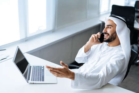 handsome man with dish dasha working in his business office of Dubai. Portraits of a successful businessman in traditional emirates white dress. Concept about middle eastern cultures, lifestyle and professional occupationsの写真素材