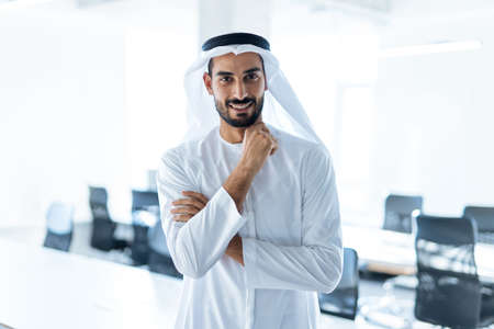 handsome man with dish dasha working in his business office of Dubai. Portraits of a successful businessman in traditional emirates white dress. Concept about middle eastern cultures, lifestyle and professional occupationsの写真素材