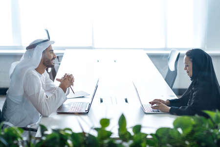 Man and woman with traditional clothes working in a business office of Dubai. Portraits of successful entrepreneurs businessman and businesswoman in formal emirates outfits. Concept about middle eastern cultures, lifestyle and professional occupationsの写真素材