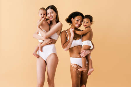 Happy women holding their babies on colored background - Young women wearing underwear taking care of their little sons - Pregnancy, motherhood, people and expectation conceptの写真素材