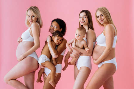 Happy pregnant women with big belly and beautiful mothers with babies in studio - Multiracial group of pregnant women and moms with childs wearing underwear - Pregnancy, motherhood, people, body positive and body acceptance conceptの写真素材