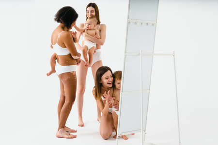 Happy multiethnic women holding their babies on colored background - Young women wearing underwear taking care of their little sons - Pregnancy, motherhood, people, body positive and body acceptance conceptsの写真素材