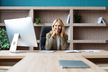 Beautiful mature adult businesswoman with elegant dress sitting at computer desk in the office and talking at mobile phone - Senior woman working at pc online, concepts about business and technologyの写真素材
