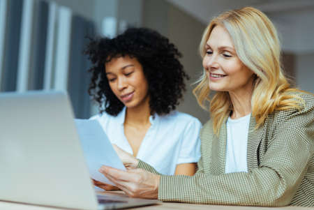 Beautiful adult businesswomen with elegant dress sitting at computer desk in the office - Senior caucasian woman and younger african american female working at laptop online, concepts about business and technologyの写真素材