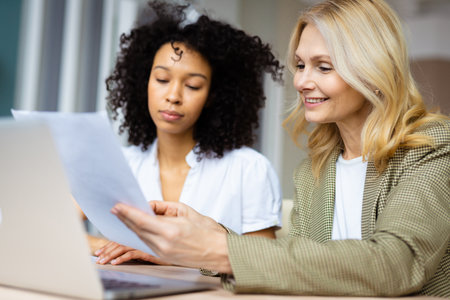 Beautiful adult businesswomen with elegant dress sitting at computer desk in the office - Senior caucasian woman and younger african american female working at laptop online, concepts about business and technologyの写真素材
