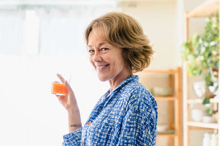 Beautiful mature senior woman at home, domestic life and leisure moments - 50-60 years old pretty female adult drinking healthy orange juice for breakfastの写真素材