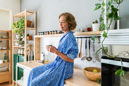 Beautiful mature senior woman at home, domestic life and leisure moments - 50-60 years old pretty female adult drinking coffee in the morning in the kitchenの写真素材