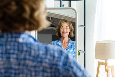 Beautiful mature senior woman at home, domestic life and leisure moments - 50-60 years old pretty female adult looking at herself in the mirrorの写真素材