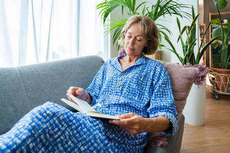 Beautiful mature senior woman at home, domestic life and leisure moments - 50-60 years old pretty female adult relaxing on the sofa and reading a bookの写真素材