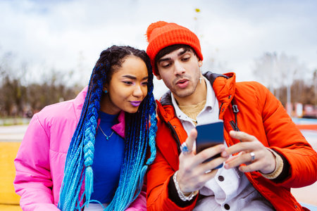 Multiracial young couple of lovers dating outdoors in winter using social media app on smartphone, wearing winter jackets and having fun - Multiethnic millennials bonding in an urban area, concepts about youth and social releationshipsの写真素材