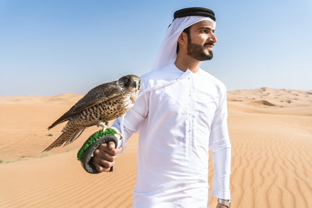 Middle-eastern man wearing traditional emirati arab kandura in the desert and holding a falcon bird...