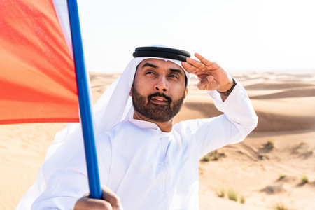 Middle-eastern man wearing traditional emirati arab kandura in the desert holding the UAE flag - Arabian muslim adult person at the sand dunes in Dubai celebrating patriotism on national dayの写真素材