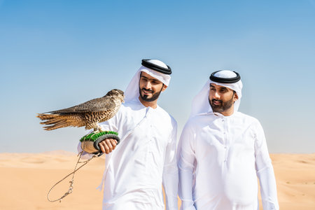 Two middle-eastern men wearing traditional emirati arab kandura bonding in the desert and holding a falcon bird - Arabian muslim friends meeting at the sand dunes in Dubaiの写真素材