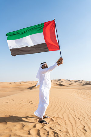 Middle-eastern man wearing traditional emirati arab kandura in the desert holding the UAE flag - Arabian muslim adult person at the sand dunes in Dubai celebrating patriotism on national dayの写真素材