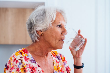 Beautiful senior old woman with gray hair drinking glass of water at home - Youthful elderly granmother spending quality time at homeの写真素材