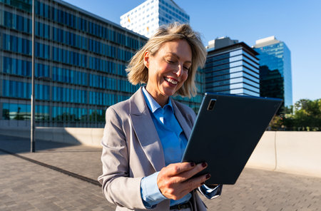 Beautiful middle aged businesswoman wearing elegant suit using tablet device - Corporate mature adult female with stylish formal attire remote working online outdoorsの写真素材