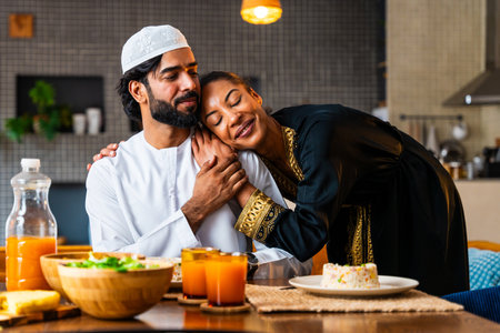 Happy beautiful young arab couple with emirati clothing having lunch together at home - Middle-eastern man and woman with traditional arabic attire eating healthy food after ramadanの写真素材