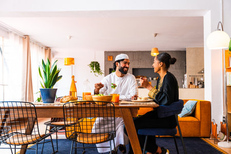 Happy beautiful young arab couple with emirati clothing having lunch together at home - Middle-eastern man and woman with traditional arabic attire eating healthy food after ramadanの写真素材