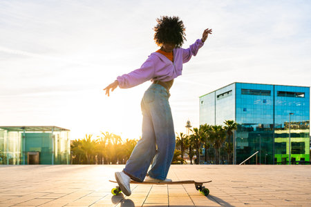 Beautiful young black woman with curly afro hair portrait outdoors - Modern cool female stylish wearing urban clothing strolling in the city dancing on longboardの写真素材