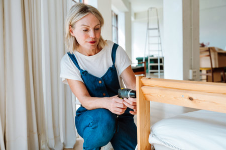Single woman relocating in a new apartment. Middle aged senior beautiful woman fixing the head of the bed with an electric screwdriver.の写真素材