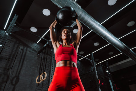 Cinematic image of a fit young woman in red sport outfit doing workout in the gym. Female athlete using medicine ball to train strength.の写真素材