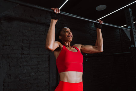Cinematic image of a fit young woman in red sport outfit doing workout in the gym. Young athlete doing chin ups at the bar.の写真素材
