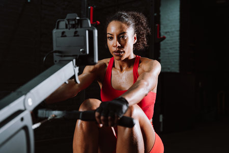 Cinematic image of a fit young woman in red sport outfit doing workout in the gym. Woman using the rowing machine.の写真素材
