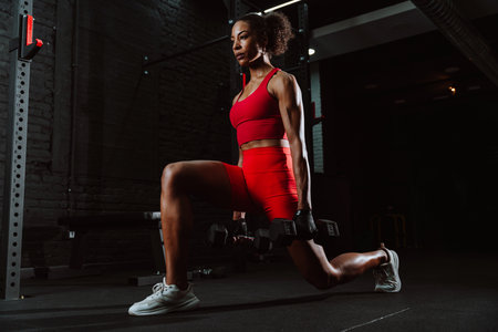 Cinematic image of a fit young woman in red sport outfit doing workout in the gym. Woman doing lunges with dumbbells.の写真素材