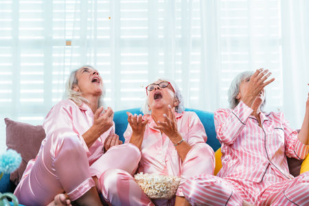 Beautiful senior women having funny pajama party at home - Group of cheerful elderly female friends eating pop-corn while watching series on tv, concepts about old people, lifestyle and quality of lifeの写真素材