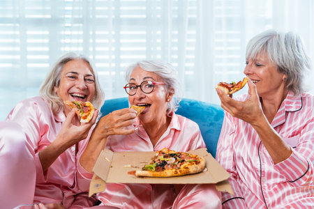 Beautiful senior women having funny pajama party at home - Group of cheerful elderly female friends having fun at home eating pizza, concepts about old people, lifestyle and quality of lifeの写真素材