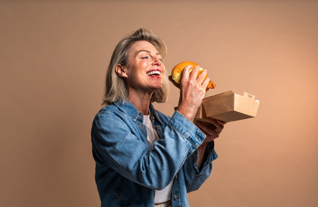 Beautiful mature woman eating tasting hamburger on colorful background - Portrait of pretty middle aged female holding fast food sandwichの写真素材