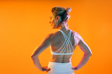 Beautiful mature sportive woman wearing sportswear doing workout - back view portrait of a strong, athletic mature woman in white sportswear, posing with hands on hips against an orange backgroundの写真素材