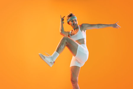 Beautiful mature sportive woman wearing sportswear doing workout - energetic senior woman dancing and exercising in a vibrant orange studio, embodying fitness and an active lifestyleの写真素材