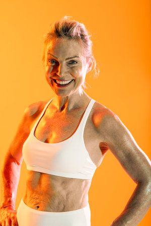 Beautiful mature sporty woman wearing sportswear doing workout - mature sportswoman in white athletic wear smiling confidently. Displaying toned abs and fit physique against vibrant orange backgrounの写真素材