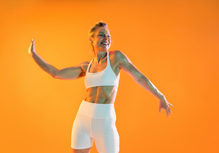 Beautiful mature sportive woman wearing sportswear doing workout - happy, sporty mature woman in white sportswear enjoying a workout on an orange background, promoting a healthy lifestyleの写真素材