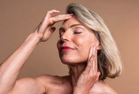 Beauty portrait of beautiful mature woman, concepts about cosmetics, skincare and wellness. Mature woman with closed eyes gently touching face, embodying graceful aging and radiant skin wellnessの写真素材