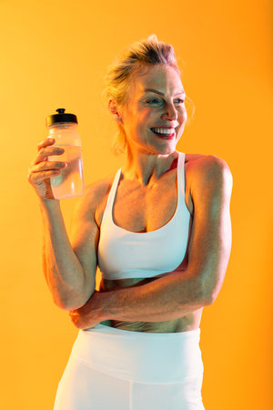 Beautiful mature sportive woman wearing sportswear doing workout - happy mature woman in white sportswear, holding a water bottle and smiling after an intense workout against a vibrant orange backdropの写真素材