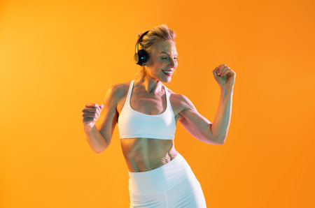 Beautiful mature sportive woman wearing sportswear doing workout - athletic mature woman wearing headphones and sportswear dances energetically and smiles against a vibrant orange backdropの写真素材