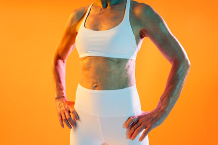 Beautiful mature sportive woman wearing sportswear doing workout - confident senior fitness woman wearing white sportswear, proudly displaying her toned body and muscles against an orange backdropの写真素材