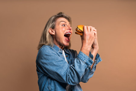 Beautiful mature woman eating tasting hamburger on colorful background - Portrait of pretty middle aged female holding fast food sandwichの写真素材