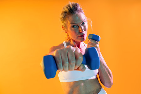 Beautiful mature sportive woman wearing sportswear doing workout - concentrated mature sportswoman lifting blue dumbbells against an orange background, exemplifying strength and commitment to fitnessの写真素材
