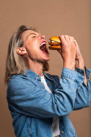 Beautiful mature woman eating tasting hamburger on colorful background - Portrait of pretty middle aged female holding fast food sandwichの写真素材