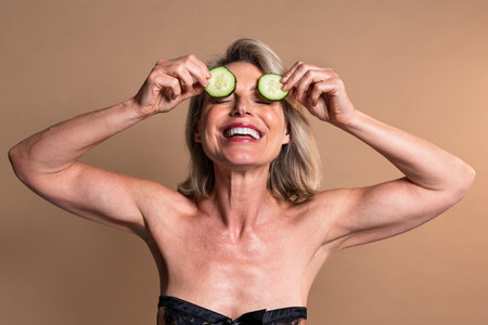 Beauty portrait of beautiful mature woman, concepts about cosmetics, skincare and wellness. Happy mature woman with bare shoulders holding cucumber slices on her eyesの写真素材