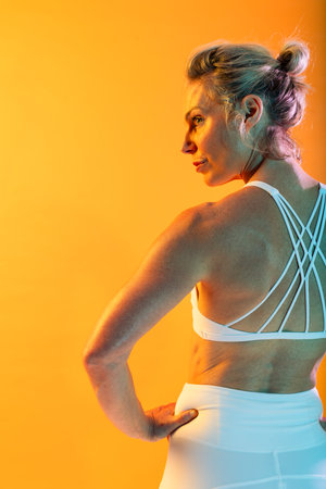 Beautiful mature sportive woman wearing sportswear doing workout - muscular mature woman in white sportswear posing with hands on hips and looking over her shoulder against an orange backgroundの写真素材
