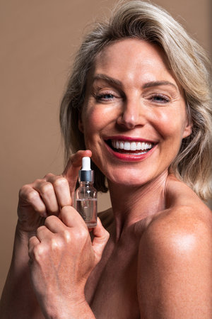 Beauty portrait of beautiful mature woman, concepts about cosmetics, skincare and wellness. Mature woman smiling while holding a bottle of essential oil serumの写真素材